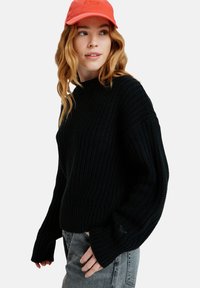 Black knitted sweater with a high neckline, relaxed silhouette, and wide cuffs, paired with a coral cap featuring embroidered text.