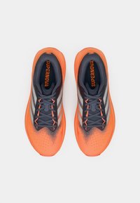 Buty sportowe w kolorze pomarańczowym i szarym, z teksturowaną cholewką, perforacjami, sznurowadłami oraz napisem "SUPERNOVA" na języku.