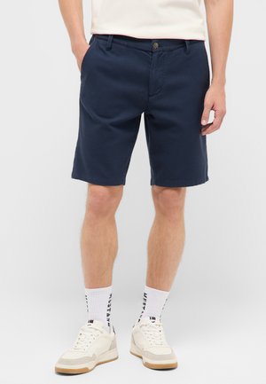 Mann trägt marineblaue knielange Shorts, weiße Socken mit schwarzem Text und weiße Sneaker mit beigen Sohlen.