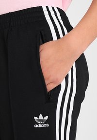 Czarne sportowe spodnie z białymi paskami, wyposażone w elastyczny pas i boczną kieszeń na zamek, ozdobione małym białym logo Adidas.