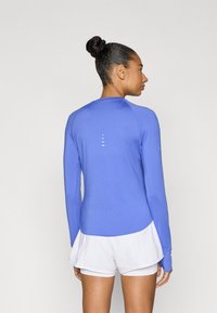 Μακρυμάνικη αθλητική μπλούζα σε φωτεινό μπλε χρώμα, κατασκευασμένη από λείο ύφασμα με κυρτό τελείωμα και διακριτικές λευκές λεπτομέρειες στην πλάτη. Συνδυάζεται με λευκά σορτς.