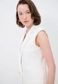 Bílý bezrukávový blazer se strukturovanými rameny, zapínáním na jeden knoflík a předními kapsami. Hladká textura a minimalistický design.