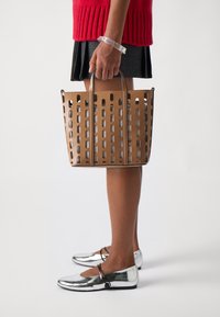 Beige geperforeerde tote bag met dubbele handvatten, met een gestructureerde vorm en een gladde textuur. Gecombineerd met metallic zilveren platte schoenen en een gebreide rode top.