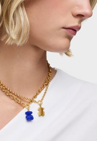 Collar de cadena dorada que presenta dos charms: un osito de peluche azul y una silueta de oso dorado, usado sobre una prenda blanca de canalé.