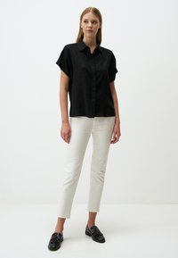 Schwarzes kurzärmeliges Button-up-Hemd aus texturiertem Stoff, kombiniert mit eng anliegenden weißen Hosen und schwarzen Slippern mit Quastenakzenten.