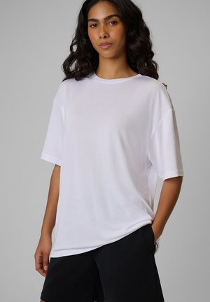 Kobieta o długich, kręconych ciemnych włosach, ubrana w luźny biały t-shirt i czarne szorty, stojąca z jedną ręką w kieszeni na szarym tle.