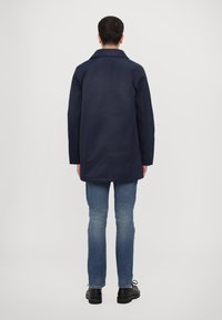 Navy blue coat with a collar, long sleeves, and a straight cut. Worn over light blue jeans and black shoes. Smooth texture.
