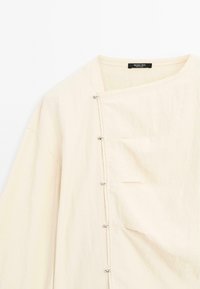 Blouse beige clair avec un design structuré, col rond, poche latérale et accents de boutons métalliques le long de la couture avant. Tissu doux et texturé.