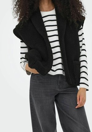 Kvinde iført sort sherpa-vest over hvid og sort stribet langærmet skjorte og mørkegrå jeans, hånd i lommen, står mod en hvid baggrund.