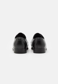 Chaussures habillées en cuir noir avec une finition brillante, bout arrondi et petit talon empilé, présentant une texture lisse et des détails de couture minimalistes.