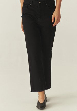 Personne portant un jean noir taille haute à jambes larges avec un ourlet brut et des chaussures noires à talons bas, debout devant un fond neutre.