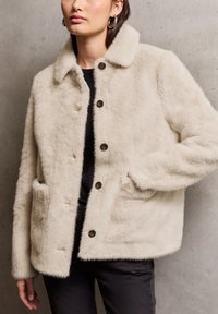 Veste en fausse fourrure crème avec un col, fermeture à boutons et deux poches avant. Elle possède une texture douce et une coupe décontractée.