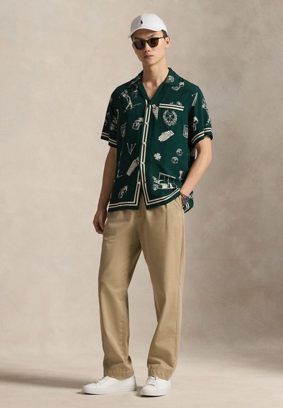 Mężczyzna w ciemnozielonej, wzorzystej koszuli z krótkim rękawem, beżowych spodniach, białych trampkach, białej czapce i okularach przeciwsłonecznych, stojący z jedną ręką w kieszeni.