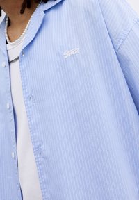 Camicia a righe blu chiaro con chiusura a bottoni, dotata di un logo ricamato sul petto sinistro e una maglietta bianca visibile sotto.