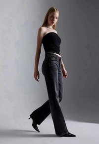 Młoda kobieta idąca, ubrana w czarny top na ramiączkach, ciemne szerokie dżinsy i czarne szpilki z wąskim noskiem, na tle szaro-zielonym gradientem.