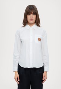 Camisa blanca de manga larga con cuello punta y dobladillo redondeado. Incluye un bolsillo en el pecho con un parche de logo marrón. Textura de tela suave.