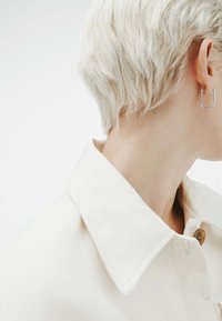 Persona con pelo corto rubio platino, vistiendo una camisa blanca con cuello y un pendiente rectangular plateado en la oreja derecha.
