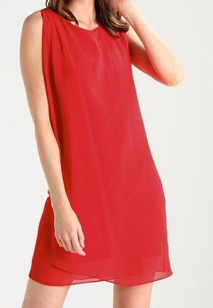 Femme portant une robe rouge sans manches, ample, avec un tissu transparent superposé, se tenant devant un fond clair uni.