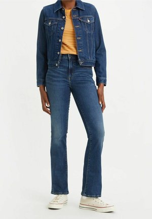 Person trägt blaue Jeansjacke, blaue Jeans mit geradem Bein, gelb gestreiftes Hemd und weiße Turnschuhe und steht vor einem schlichten Hintergrund.