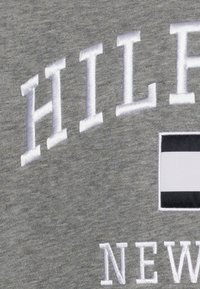 Felpa grigia con ricamo del testo "HILFIGER" in bianco, con un logo rettangolare in bianco e nero. Morbido tessuto di cotone.
