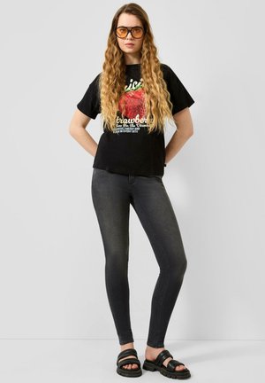 Frau mit langen, welligen Haaren, die orange getönte Brillen, ein schwarzes T-Shirt mit Erdbeergrafik, dunkle Skinny-Jeans und schwarze Sandalen trägt, steht vor einer grauen Wand.