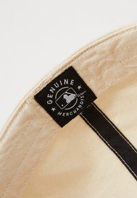 Crème kleurige pet met een zwart rechthoekig label binnenin, met witte tekst die "ECHTE MERCHANDISE" zegt en een logo-embleem.