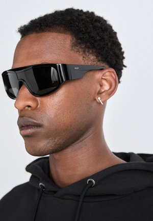 Black oversized sunglasses with angular design, featuring a glossy finish and gold "RSF" branding on the temples. Worn with a black hoodie.
