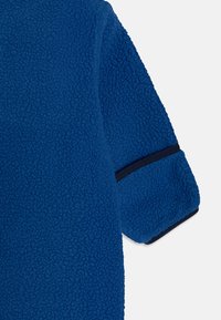 Tecido de polar azul com um acabamento texturizado; apresenta uma faixa azul marinho na bainha. O design inclui uma forma de manga larga e descontraída.