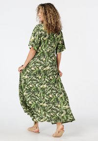 Maxi-jurk van groene stof met een tropisch bladeren- en ananasprint. Ruime pasvorm met korte mouwen en een ronde halslijn; gedragen met tan sandalen.
