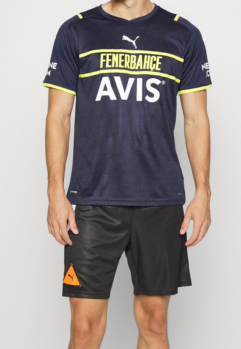 Tummansininen urheilu paita, jossa on keltaiset raidat, sisältäen Fenerbahçe- ja AVIS-logot; yhdistetty mustiin tekstuurihousuihin ja oranssiin PUMAn logoon.