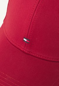 Red baseball cap made of textured fabric with curved visor. Features a small logo with black, white, and red stripes on the side.
