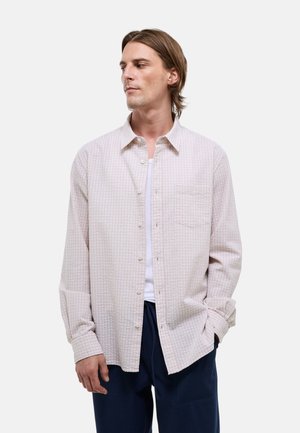 Homme aux cheveux mi-longs portant une chemise claire à carreaux boutonnée sur un t-shirt blanc et un pantalon foncé, debout avec une main dans la poche.