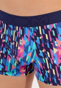 Swim shorts with a navy blue elastic waistband, featuring a geometric pattern in pink, green, and blue tones on a dark background.