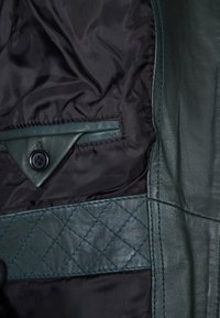 Zelená kožená bunda uvnitř s černým nylonovým podšívkou; obsahuje trojúhelníkovou koženou kapsu s knoflíkem a prošívan�ý kožený detail.