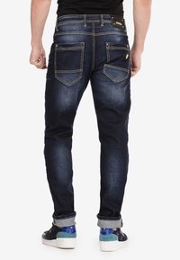 Jeans de mezclilla con un lavadado oscuro, con costuras amarillas, dos bolsillos traseros con solapas puntiagudas y dobladillos doblados. Llevados con zapatillas azules y blancas.