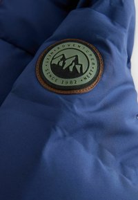 Blauer Stoff mit einem runden Patch, der Bergsilhouetten zeigt, Text "Abenteurer seit 1982 Nächster" und einem braunen Rand.