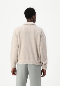 Beige fleece jas met een kraag, lange mouwen en knoopmanchetten. Zachte textuur en relaxte pasvorm. Gecombineerd met lichtgrijze broeken.