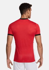 Maglia sportiva rossa a maniche corte, con dettagli neri sul colletto e sui polsini, caratterizzata da una texture a rete e un design aderente. Vista posteriore mostrata.