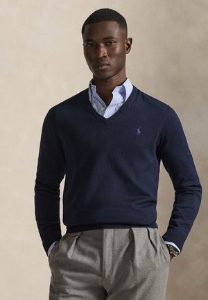 Homme portant un pull bleu marine à col en V par-dessus une chemise rayée bleue et un pantalon plissé gris, debout les mains dans les poches devant un fond uni.