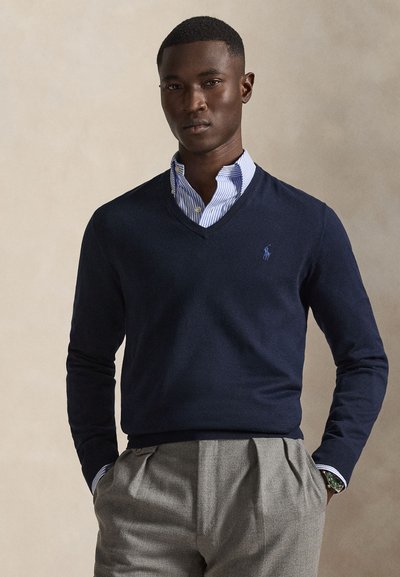Uomo con maglione blu navy a V sopra a una camicia a righe blu e pantaloni grigi plissettati, in piedi con le mani nelle tasche davanti a uno sfondo semplice.