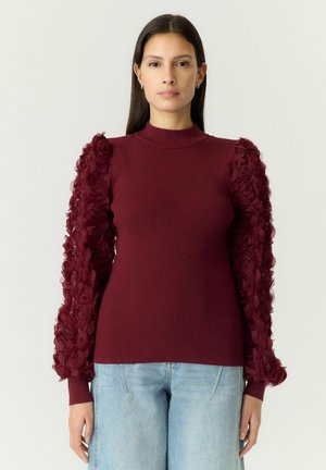 Mulher vestindo um suéter canelado cor bordô com mangas bufantes florais texturizadas e jeans azul claro, em pé contra um fundo claro.