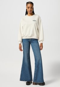 Cream-colored sweatshirt with ribbed cuffs, featuring "Wrangler 1947" embroidered in green. Paired with flared blue denim jeans.