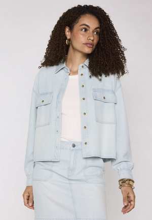 Vrouw met krullend haar die een licht denimjasje en bijpassende rok draagt met een wit topje, gouden oorringen en armbanden, zijwaarts kijkend.