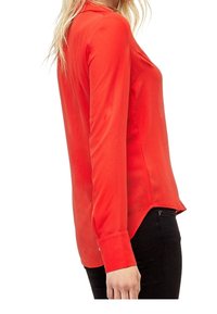 Blouse rouge à manches longues avec une texture lisse, dotée d'un col rond et d'un ourlet asymétrique, associée à un pantalon noir ajusté.