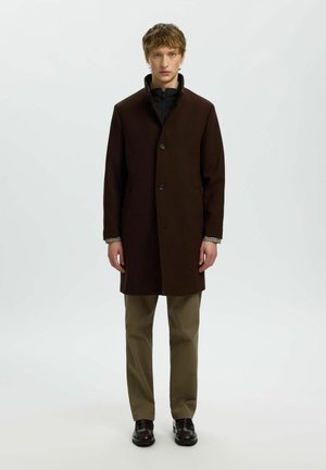 Jeune homme debout, portant un manteau marron foncé boutonné sur une chemise noire, un pantalon beige et des chaussures foncées sur un fond clair uni.