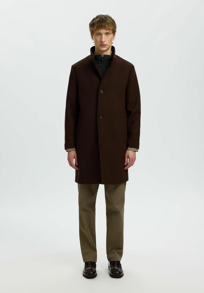 Jeune homme debout, portant un manteau marron foncé boutonné sur une chemise noire, un pantalon beige et des chaussures foncées sur un fond clair uni.