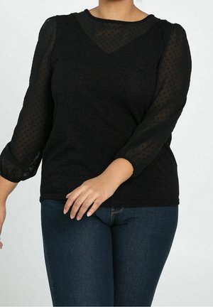 Vrouw die een zwart lang shirt draagt met doorschijnende polkadot mouwen en donkerblauwe spijkerbroek, afgebeeld van nek tot heupen.