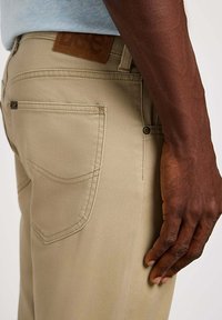 Beige katoenen broeken met een slim fit, voorzien van een leren taillebandpatch en zichtbare achterzakken, met stikseldetails en metalen hardware.