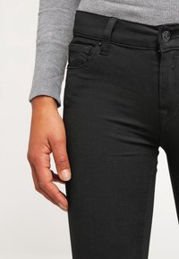 Jeans negros ajustados con una textura suave, que cuentan con cierre de botón y un bolsillo lateral. Se ve una camiseta de manga larga gris que acompaña.