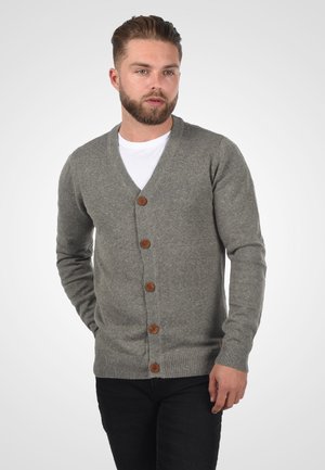 Homme barbu portant un cardigan gris boutonné sur une chemise blanche et un pantalon noir, debout devant un fond clair uni.